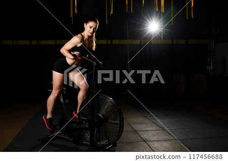 A young muscular woman is doing hard bike crossfit training in the gym. 117456688