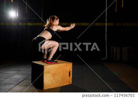 Athletic woman in sportswear jumping on crossfit box in fitness gym 117456701