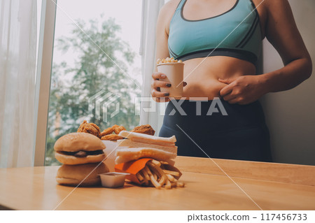 To eat or not. Cheerless plump woman looking at the burger while thinking to eat it or not To eat or not. Cheerless plump woman looking at the burger while thinking to eat it or not 117456733