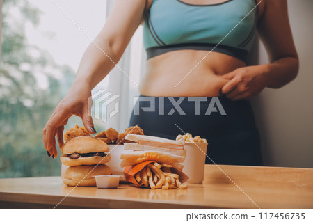 To eat or not. Cheerless plump woman looking at the burger while thinking to eat it or not To eat or not. Cheerless plump woman looking at the burger while thinking to eat it or not 117456735