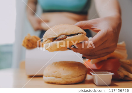 To eat or not. Cheerless plump woman looking at the burger while thinking to eat it or not To eat or not. Cheerless plump woman looking at the burger while thinking to eat it or not 117456736