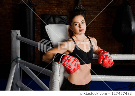Fit strong brunette woman in sportswear top and shorts in boxing gloves in the ring looks at the camera Fit strong brunette woman in sportswear top and shorts in boxing gloves in the ring looks at the camera 117457482