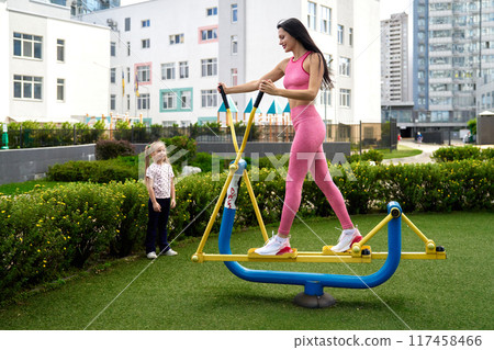 Woman exercising on outdoor fitness equipment while a child watches in a city park during daytime 117458466
