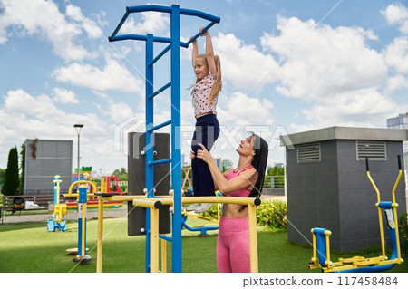 Child practicing pull-ups with adult supervision in a park gym on a sunny day 117458484