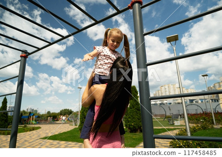 Mother lifts her daughter in a sunny playground on a summer afternoon 117458485