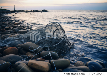 Empty fishing net resting on pebbled shoreline with calm waters at dusk Empty fishing net resting on pebbled shoreline with calm waters at dusk 117458549