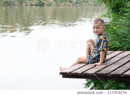 A small handsome boy, with emotional facial expressions, 4 years old, sits on a wooden bridge by the river, against the backdrop of green plants and breathes fresh air. Relax. Place for text. 117459254