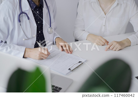 Doctor and a patient. The female physician, wearing a white medical coat over a dark blue dotted blouse, using clipboard and making notes during a consultation in the clinic. Medicine concept 117460042