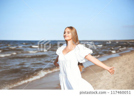Happy smiling beautiful woman is on the ocean beach in a white summer dress, open arms 117460051