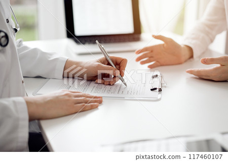 Doctor and patient sitting and discussing something near each other at the white desk in clinic. Female physician is listening filling up a records form. Medicine concept 117460107
