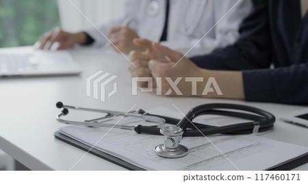 Stethoscope over a clipboard with medical papers is lying on the table near doctor and patient discussing health while sitting in clinic. Medicine and science Stethoscope over a clipboard with medical papers is lying on the table near doctor and patient discussing health while sitting in clinic. Medicine and science 117460171
