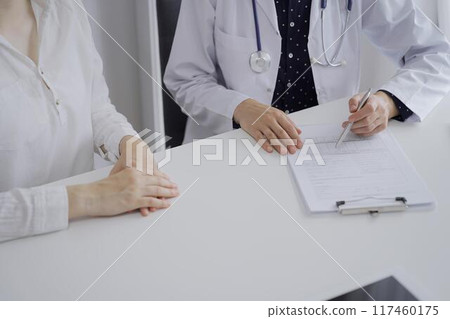 Doctor and a patient. The female physician, wearing a white medical coat over a dark blue dotted blouse, using clipboard and making notes during a consultation in the clinic, close up. Medicine Doctor and a patient. The female physician, wearing a white medical coat over a dark blue dotted blouse, using clipboard and making notes during a consultation in the clinic, close up. Medicine 117460175