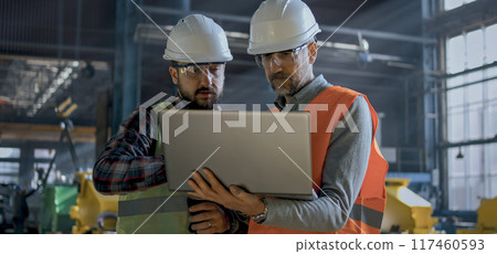 Two industry technicians stand in manufacturing factory, work on laptop Two industry technicians stand in manufacturing factory, work on laptop 117460593