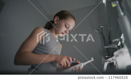 Young girl looks at mirror in bathroom, brushes her teeth Young girl looks at mirror in bathroom, brushes her teeth 117460826