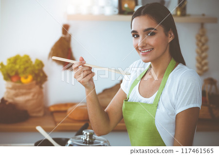 An attractive young dark-haired woman tasting ready hot meal with a wooden spoon while standing and smiling in sunny kitchen. Cooking and householding concepts An attractive young dark-haired woman tasting ready hot meal with a wooden spoon while standing and smiling in sunny kitchen. Cooking and householding concepts 117461565