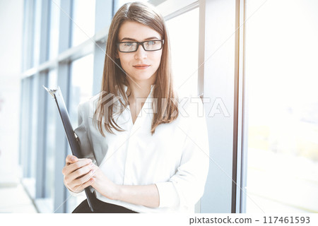 Beautiful female specialist with clipboard standing in sunny office and smiling charmingly. Business people concept Beautiful female specialist with clipboard standing in sunny office and smiling charmingly. Business people concept 117461593