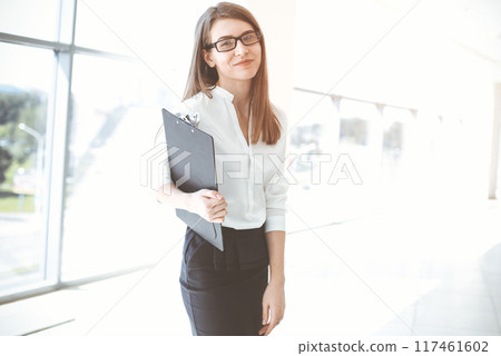 Beautiful female specialist with clipboard standing in sunny office and smiling charmingly. Business people concept 117461602