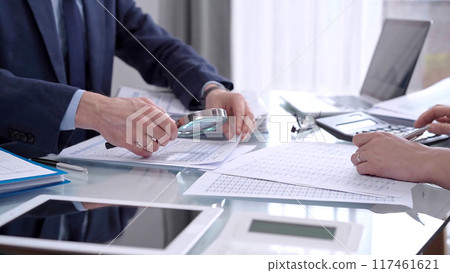 Business people wearing blue suits are reviewing financial data in office. Male auditor is using a magnifying glass. Audit and taxes concept 117461621