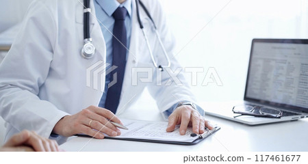 Doctor and a patient. The physician, wearing a white medical coat over a blue shirt and tie, gesturing with his hands during a consultation in the clinic. Medicine concept Doctor and a patient. The physician, wearing a white medical coat over a blue shirt and tie, gesturing with his hands during a consultation in the clinic. Medicine concept 117461677