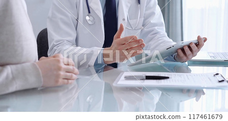 Doctor is consulting patient in modern clinic. Healthcare professional discusses medical information with a woman, using a tablet computer. Medicine concept 117461679