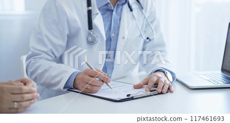 A doctor and a patient. The physician, wearing a white medical coat over a blue shirt, is filling out a medical record form during a consultation in the clinic. Medical service A doctor and a patient. The physician, wearing a white medical coat over a blue shirt, is filling out a medical record form during a consultation in the clinic. Medical service 117461693
