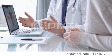 Doctor is consulting patient in modern fair clinic. Healthcare professional discusses medical information with a woman, gesturing above the laptop computer on the glass desk. Medicine 117461709