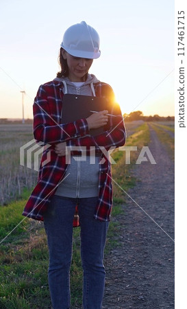 Woman engineer is standing straight and taking notes with a clipboard in a field with wind turbines, as the sun sets. Clean energy and engineering concept Woman engineer is standing straight and taking notes with a clipboard in a field with wind turbines, as the sun sets. Clean energy and engineering concept 117461715