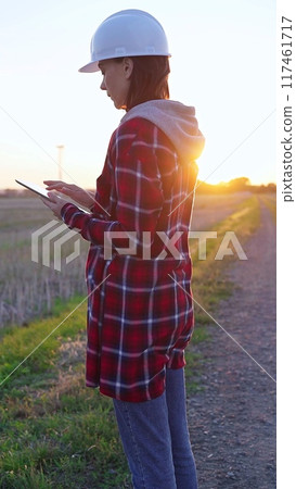 Woman engineer wearing a white protective helmet is taking notes with a clipboard in a field with wind turbines, as the sun sets. Clean energy and engineering concept 117461717