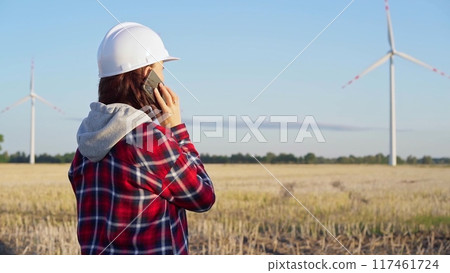 Woman engineer, wearing a white protective helmet, is talking on a smartphone in a field with wind turbines as the sun sets. Concept of clean energy and engineering audit 117461724