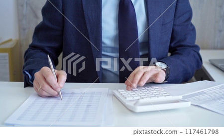 Businessman accountant with blue suit is using white calculator and taking notes with a silver pen. Taxes and audit concept Businessman accountant with blue suit is using white calculator and taking notes with a silver pen. Taxes and audit concept 117461798