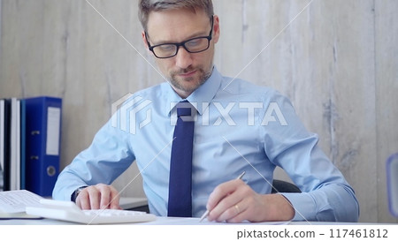 Male accountant with blue t-shirt concentrating while calculating costs and taking notes at his desk. Taxes, audit in business 117461812