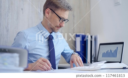 Male accountant with blue t-shirt concentrating while calculating costs and taking notes at his desk. Taxes, audit in business Male accountant with blue t-shirt concentrating while calculating costs and taking notes at his desk. Taxes, audit in business 117461813
