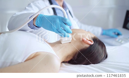 Doctor man wearing blue medical gloves is using ultrasound equipment on neck of female patient lying down for a medical examination 117461835