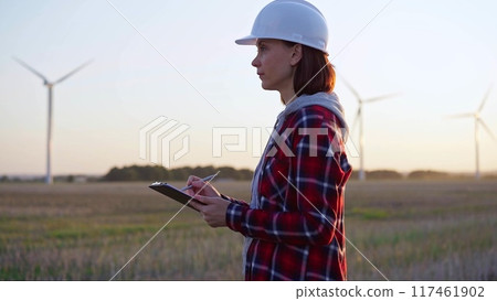 Adult woman engineer wearing white cask is taking notes on a clipboard on a field with wind turbines Adult woman engineer wearing white cask is taking notes on a clipboard on a field with wind turbines 117461902