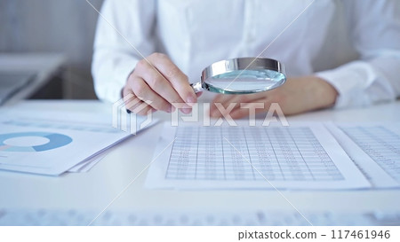Businesswoman with white blues is analyzing financial data on a spreadsheet using a magnifying glass. Audit and taxes 117461946