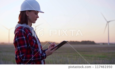 Female engineer taking notes on a clipboard on a field with wind turbines, as the sun sets. Concept of clean energy and engineering audit Female engineer taking notes on a clipboard on a field with wind turbines, as the sun sets. Concept of clean energy and engineering audit 117461950