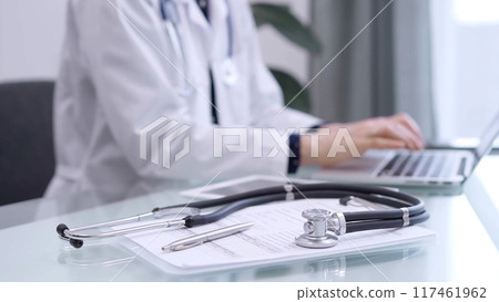 Clipboard with medical records and stethoscope are lying on the glass table while doctor woman is using tablet computer on the glass table. Medicine concept 117461962