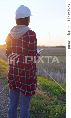 Woman engineer wearing a white protective helmet is taking notes with a clipboard in a field with wind turbines, as the sun sets. Clean energy and engineering concept 117461973
