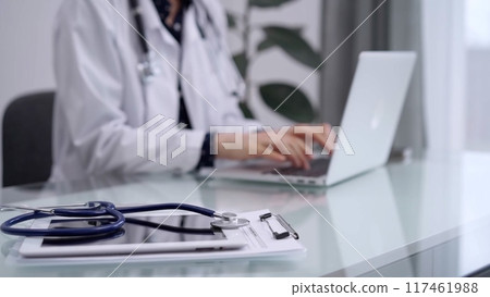 Tablet computer and blue stethoscope are on the glass table while doctor woman is using laptop computer on the glass table. Medicine concept 117461988