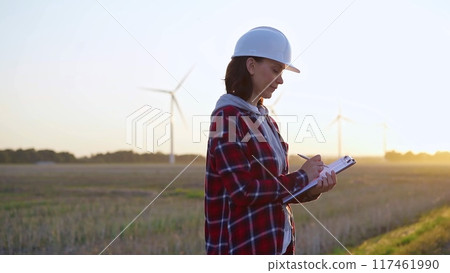 Adult woman engineer taking notes on a clipboard on a field with wind turbines, as the sun sets Adult woman engineer taking notes on a clipboard on a field with wind turbines, as the sun sets 117461990