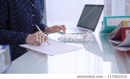 A female accountant is using a laptop computer and calculator to calculate taxes at a glass desk in the office 117462010