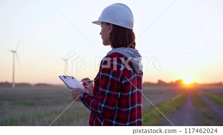Woman engineer is taking notes on a clipboard on a field with wind turbines, as the sun sets in evening. Clean energy and engineering 117462024
