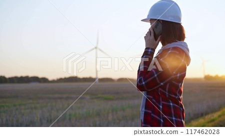 Woman engineer talking on the phone in a field with wind turbines, as the sun sets. Concept of clean energy and engineering 117462028