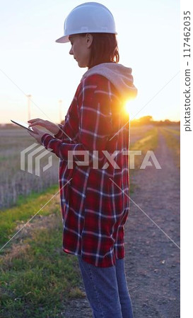 Woman engineer wearing a white protective helmet is taking notes with a clipboard in a field with wind turbines, as the sun sets. Clean energy and engineering concept Woman engineer wearing a white protective helmet is taking notes with a clipboard in a field with wind turbines, as the sun sets. Clean energy and engineering concept 117462035