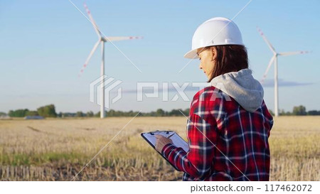 Woman engineer wearing a white protective helmet is taking notes with a clipboard in a field with wind turbines, as the sun sets. Clean energy and engineering concept 117462072