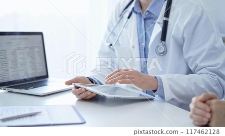 Doctor and a patient. The physician, wearing a white medical coat over a blue shirt, is using tablet computer during a consultation in the clinic. Medicine and science 117462128