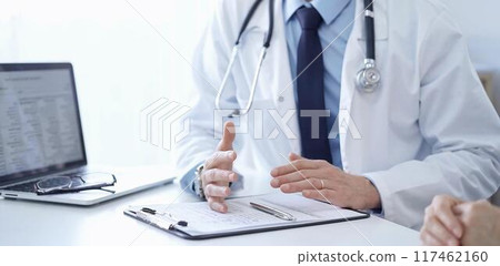 Doctor and a patient. The physician, wearing a white medical coat over a blue shirt and tie, gesturing with his hands during a consultation in the clinic. Medicine concept Doctor and a patient. The physician, wearing a white medical coat over a blue shirt and tie, gesturing with his hands during a consultation in the clinic. Medicine concept 117462160