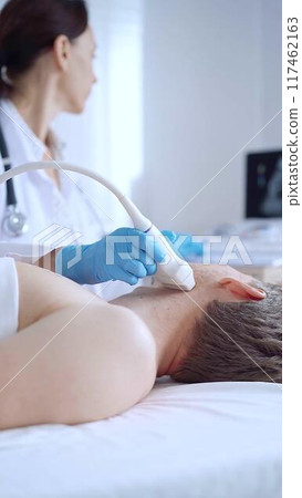Doctor woman wearing blue medical gloves performing neck ultrasound exam on male patient. Medicine service concept Doctor woman wearing blue medical gloves performing neck ultrasound exam on male patient. Medicine service concept 117462163