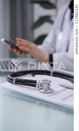 Stethoscope and medication history record form are on the glass table while doctor woman is using smartphone. Medicine theme 117462181