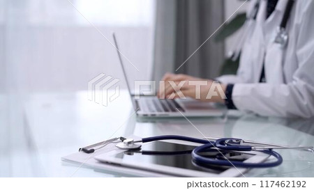 Tablet computer and blue stethoscope are on the glass table while doctor woman is using laptop computer on the glass table. Medicine concept Tablet computer and blue stethoscope are on the glass table while doctor woman is using laptop computer on the glass table. Medicine concept 117462192